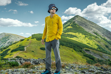 Young man standing confidently on a mountain ridge, wearing sunglasses, a yellow hiking shirt, and a sun hat, with scenic green peaks and a blue sky in the background