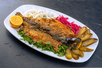 Whole cooked fish on a white oval plate, garnished with fresh parsley. Accompanied by half a lemon, sliced onions, pickled red cabbage, and cucumbers. Vibrant and visually appealing.