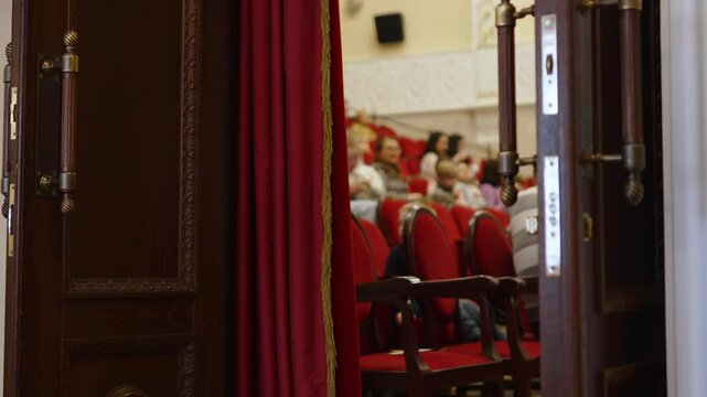 View from behind half-open doors onto blurred red theatre hall. Audience waiting for performance to begin, doorway shot. Intermission after first act, spectators rest.