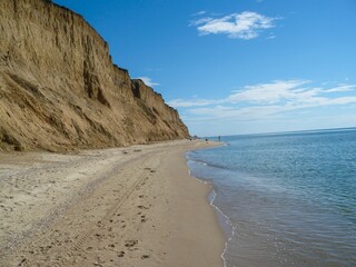 A beach with a cliff in the background. The beach is empty and the water is calm. The sky is clear and the sun is shining.