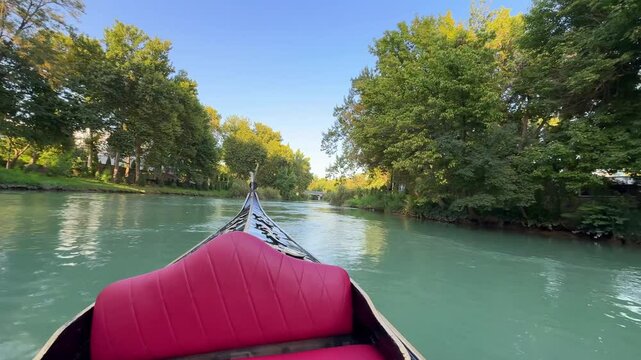 Peaceful gondola ride on clear waters surrounded by lush greenery. POV of vintage boat trip floating peacefully down a stream at sunset. Sailboat excursion across the canals of Tashkent, Uzbekistan.