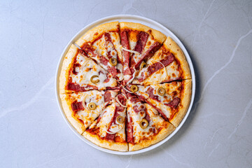 Whole pizza with slices of pepperoni, green olives, and melted cheese. Placed on a white plate, set on a light gray, marble-like surface. Freshly baked with a golden-brown crust.
