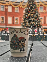 Cup with a bear on it sits on a table next to a Christmas tree. The cup is decorated with Christmas decorations and the bear is wearing a red hat. The scene is festive and cozy, with the cup