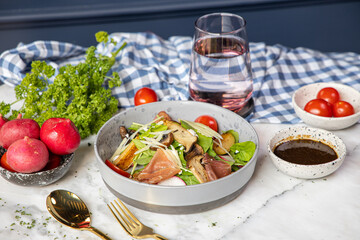 A vibrant salad bowl with fresh greens, tomatoes, and sliced meat, accompanied by radishes, dressing, and a glass of water, set against a checkered cloth backdrop.