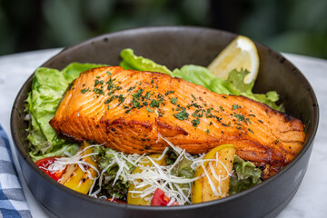 A beautifully plated salmon fillet atop fresh greens and colorful vegetables, garnished with herbs and a slice of lemon.