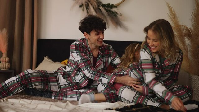 Joyful parents and their little daughter in pajamas tickle each other on the bed