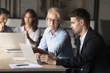 Fototapeta premium Elderly woman explain task on laptop to male colleague. Coach provide explanation about data, report, presentation to apprentice during meeting in office, discussing strategy related to joint project