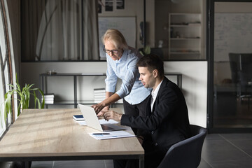 Older mentor helps with task to young staff member, showing new project details, explain corporate...