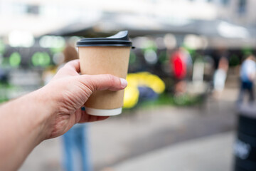 A hand holding a disposable coffee cup in a vibrant outdoor cafe setting. Blurred people and tables in the background create a lively atmosphere, perfect for leisure and socializing.