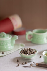 A ceramic teapot, cups, and a bowl with rolled Chinese jasmine green tea leaves are arranged on a textured surface. A mat and metal tongs for lifting hot cups complete the tea preparation scene.