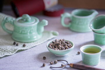 A ceramic teapot, cups, and a bowl with rolled Chinese jasmine green tea leaves are arranged on a textured surface. A mat and metal tongs for lifting hot cups complete the tea preparation scene.