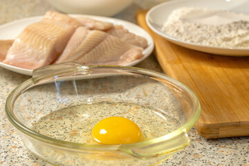 Raw chicken egg is broken into a glass plate for making a liaison. In the background are pieces of fresh fish and a bowl of flour. Close-up. Cooking at home