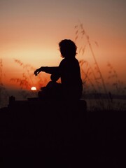 The silhouette of a girl meditating among ears of wheat on top of a hill with the sea, the sunset and the sky in the background. 