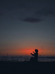 The silhouette of a girl meditating among ears of wheat on top of a hill with the sea, the sunset and the sky in the background. 