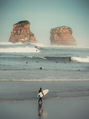 A person with a surfboard on the beach shore watching the surfers in the waves with the rocks and cliff in the background. 