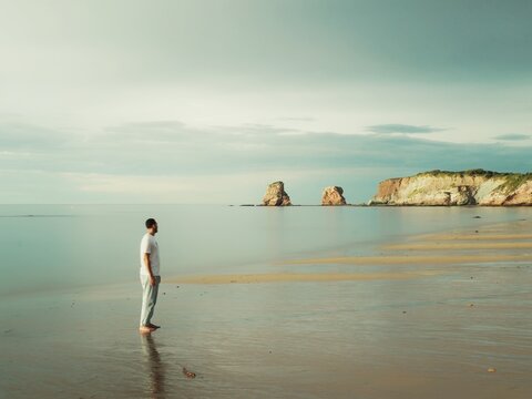 A lonely man on the shore of the beach looks out over the cliff with the silky water and sunny sky in the background. 