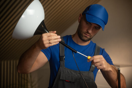 A man is fixing a lamp in a warm, inviting room filled with natural light. He adjusts the bulb carefully while wearing casual work attire. Tools are near him, indicating a DIY project.