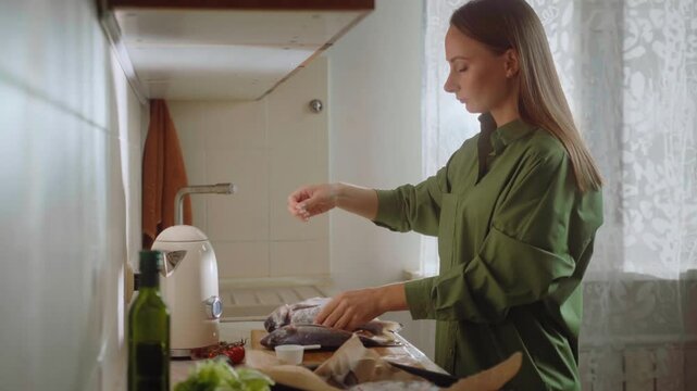 In a comfortable kitchen, a person prepares fish by carefully salting it as part of the cooking process. Fresh ingredients create a welcoming atmosphere for home cooking.