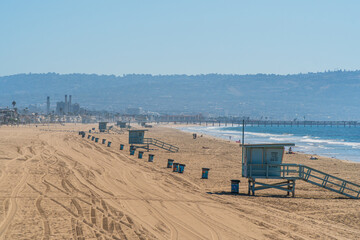 Californian beach near Los Angeles with typical lifeguard huts and garbage bins aligned on the sand, and El Segundo power plant in the distance