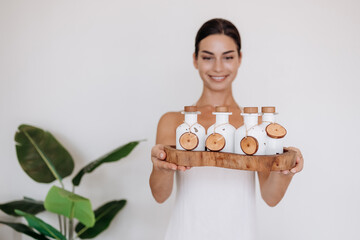 Attractive brunette smiling woman holding a bottle of cosmetic product dressed in boho white clothes. Cosmetic beauty product branding mockup. Copy space. Organic, natural product for skincare and spa