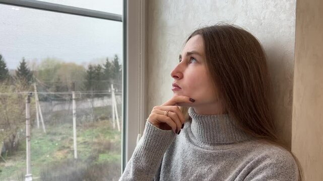 Young woman is leaning against a wall and holding her chin, deep in thought looking out a window