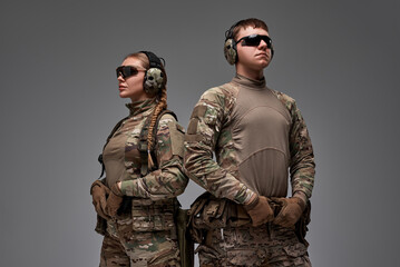A male and female tactical duo stand back-to-back in military camouflage, exuding teamwork and strength.Wearing protective gear, gloves, and headphones, they embody readiness and modern combat skills.