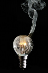 gebrochene Glühlampe mit durchbrennender  Glühwendel, und rauchendem Wolframdraht vor schwarzen Hintergrund und komplett sichtbaren Lampengewinde E29