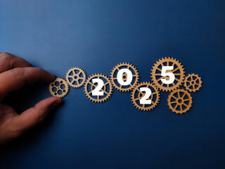2025 written on a wooden gear with someone hand holding the gear on a blue background.Business new...