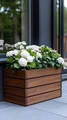 A stylish wooden planter box showcases vibrant flowers, including lavender and daisies, positioned beside a contemporary house on a bright, sunny day.