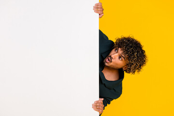 Young man with curly hair popping out from behind white board against yellow background looking...