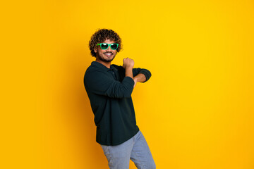 Young man in black shirt and jeans posing playfully with green sunglasses against a bright yellow background