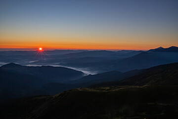 The sun rises over the mountains, fog covers the valleys, dark silhouettes of the mountains around