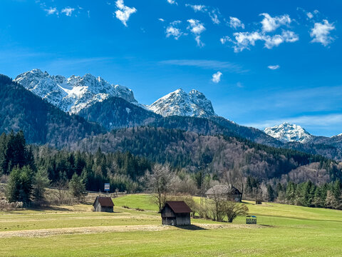 Wooden barns on alpine meadow with panoramic view of snow capped mountain peaks of Julian Alps seen from Ugovizza village, Friuli Venezia Giulia, Italy. Wanderlust in alpine wilderness in early spring