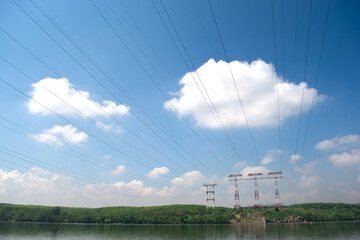 High-voltage power lines stretch across a serene landscape with a clear blue sky and fluffy white clouds, reflecting on a calm lake. Ideal for energy, nature, and infrastructure themes