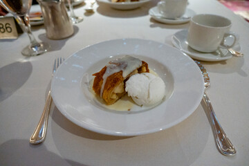 A plate of apple strudel topped with vanilla sauce, served alongside a scoop of vanilla ice cream on a white table setting.