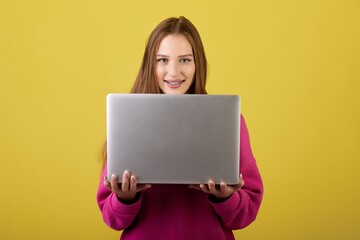 Naklejka premium Joyful woman with beautiful brown hair using a sleek silver laptop on a vibrant yellow background