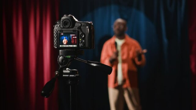 Shot with changing focus of digital camera on tripod filming performance of African American male stand-up comedian joking on small stage with red and blue curtains, humorous live show