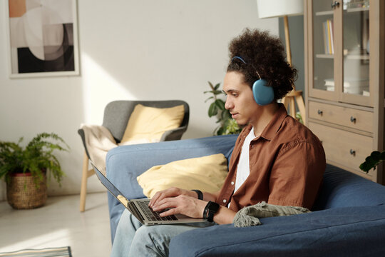 Side view of young male web designer in headphones typing and looking at laptop screen while creating new website