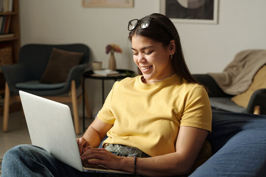 Young cheerful woman in casualwear typing on laptop keyboard while communicating with friend in video chat