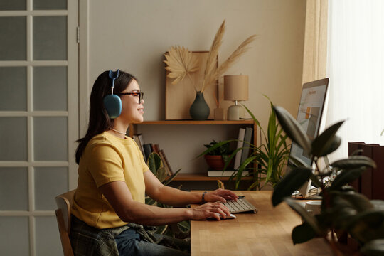 Young smiling woman in casual apparel and headphones sitting by workplace in front of desktop computer
