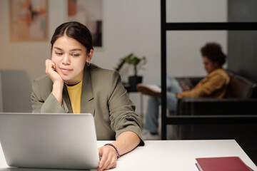 Young smiling female office worker looking at laptop screen while watching online video in coworking space