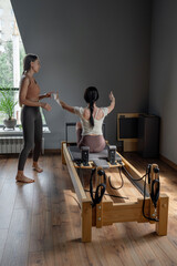 Fototapeta premium Woman training pilates on the reformer bed with trainer. Reformer pilates studio machine for fitness workouts in gym. Fit, healthy and strong authentical body. Fitness concept