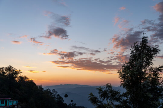 Sunset sky with vibrant orange and blue hues, scattered pink clouds and silhouetted trees against a backdrop of distant hills.A serene and tranquil village landscape near Almora in Uttarakhand, India.