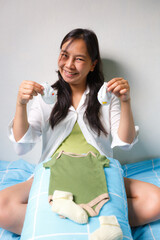 smiling pregnant woman sits on bed holding baby socks, with green baby onesie and yellow socks placed on pillow in front of her, expressing joy and anticipation