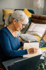 Focused senior woman working from home taking notes in notebook