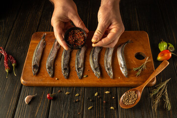 Hands skillfully sprinkle aromatic spices over a line of fresh fish on a wooden cutting board....