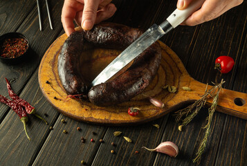 Hands carefully slice through a dark, handmade sausage on a rustic wooden board surrounded by...