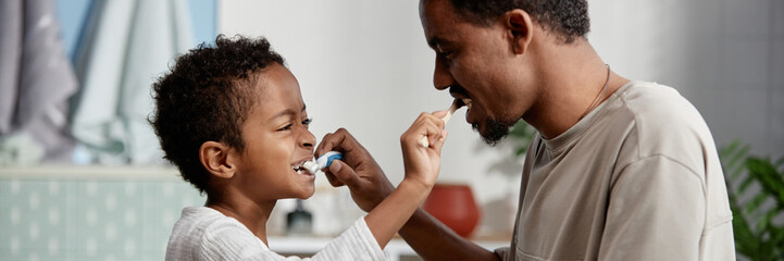 Header Image of Father Teaching Son Dental Hygiene while Black Boy Brushing Dads Teeth in Bathroom