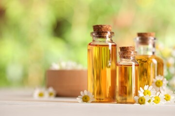 Bottles of essential oil and chamomile flowers on white wooden table against blurred background, closeup. Space for text