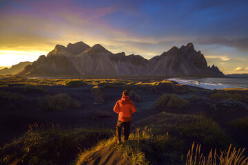 Fototapeta premium A hiker in a red rain jacket stands on a grassy sand dune at sunset, admiring Vestrahorn and Klifatindur in Stokksnes, Iceland.